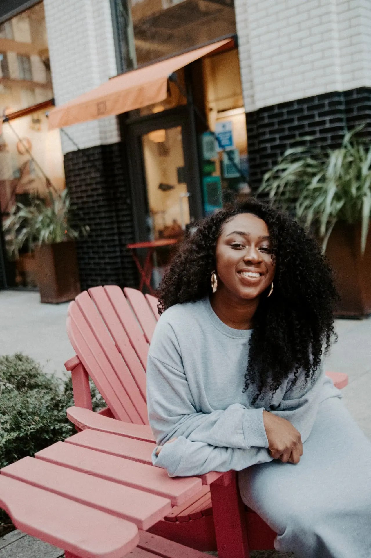 Marcelle Adrianna smiling, seated on a pink chair outside a London cafe in a grey sweatshirt set.