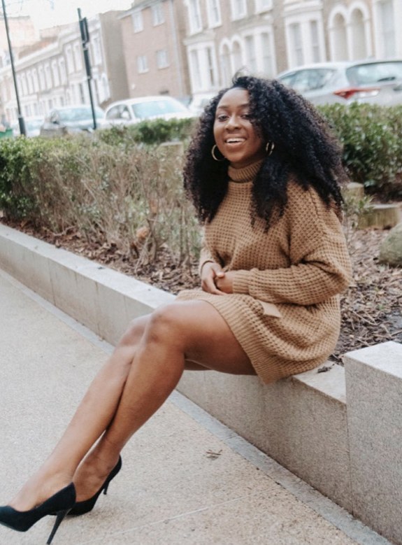 Marcelle Adrianna seated on a low wall in a neutral knit dress and black heels.