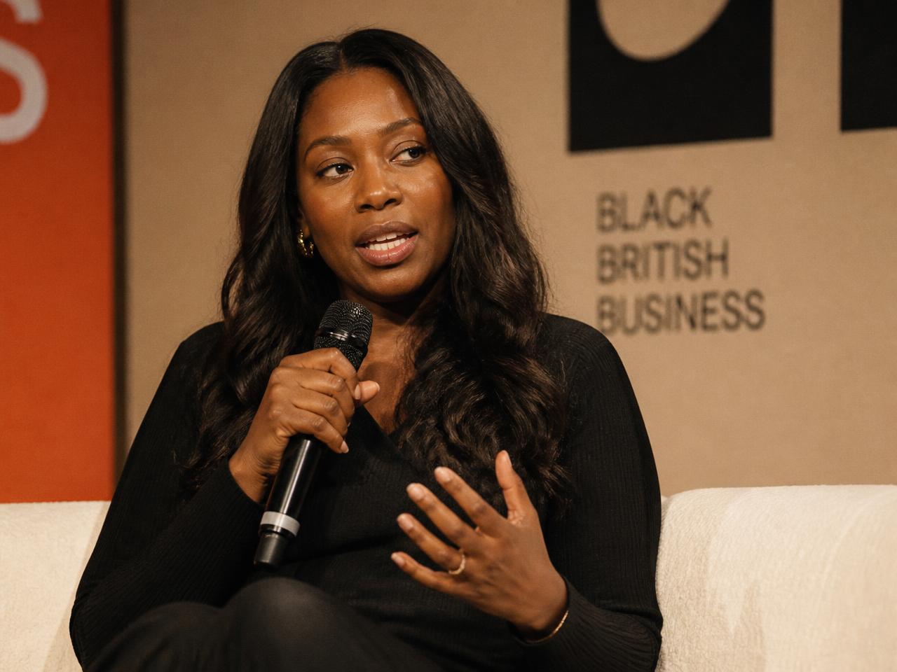 Marcelle Adrianna speaking on a panel stage, microphone in hand, warm stage light.